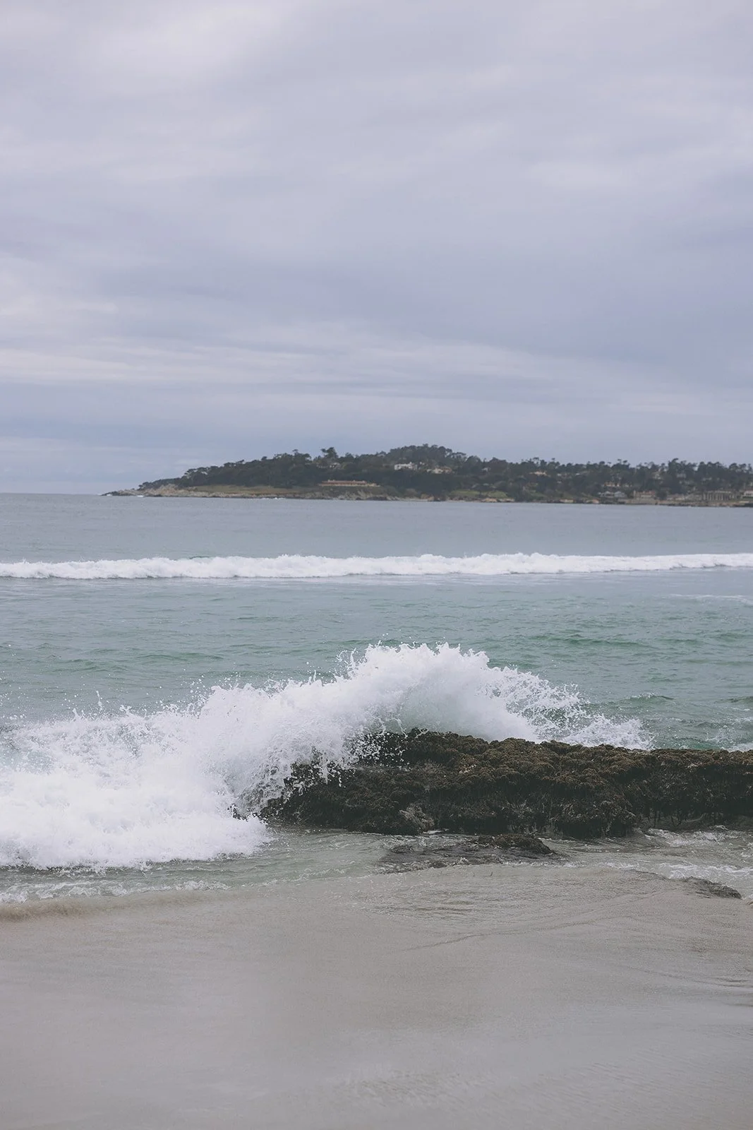 Ocean waves crashing on a rocky shore with an island in the background under a cloudy sky.