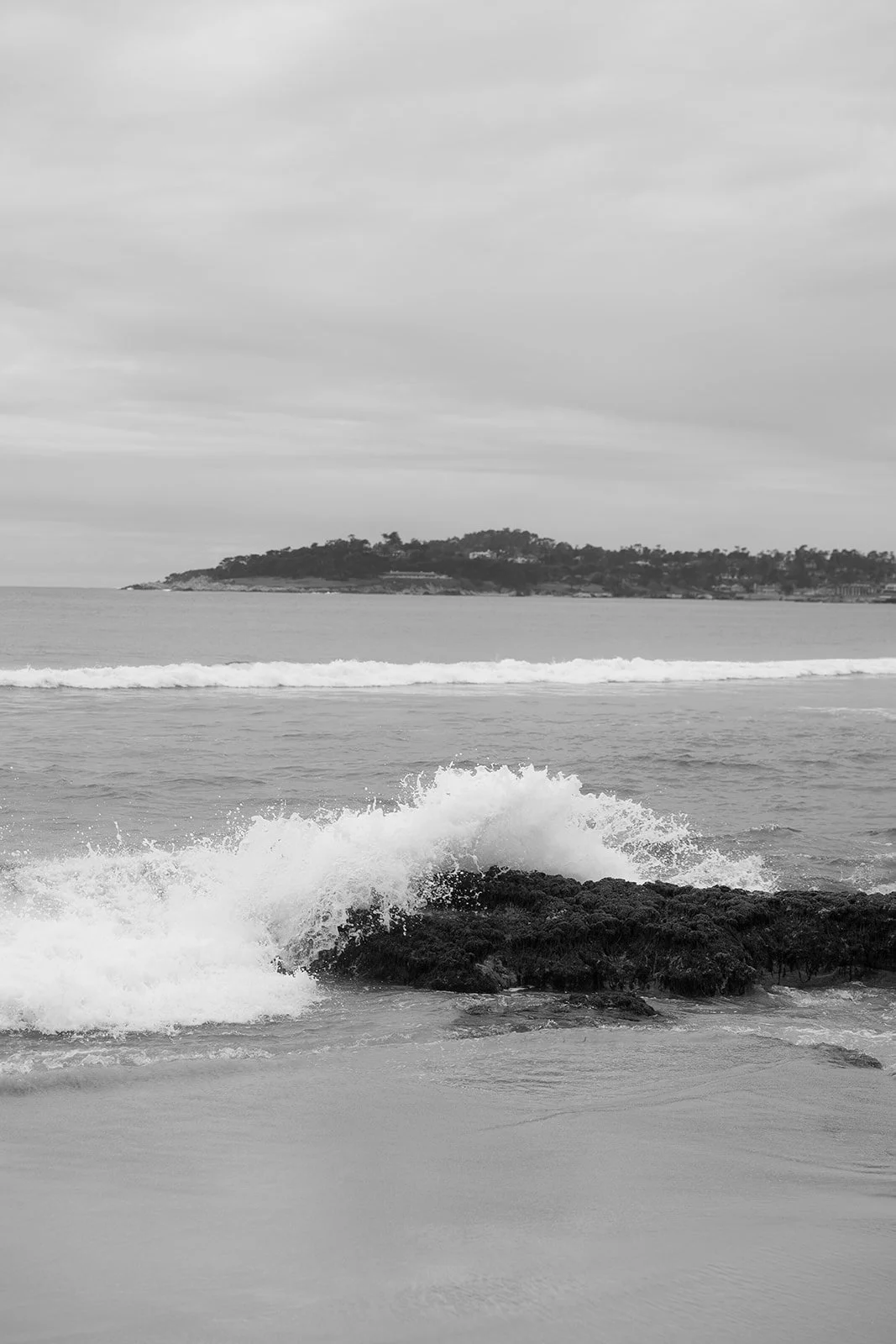 Black and white photo of waves crashing against rocks on a beach, with an island in the background.