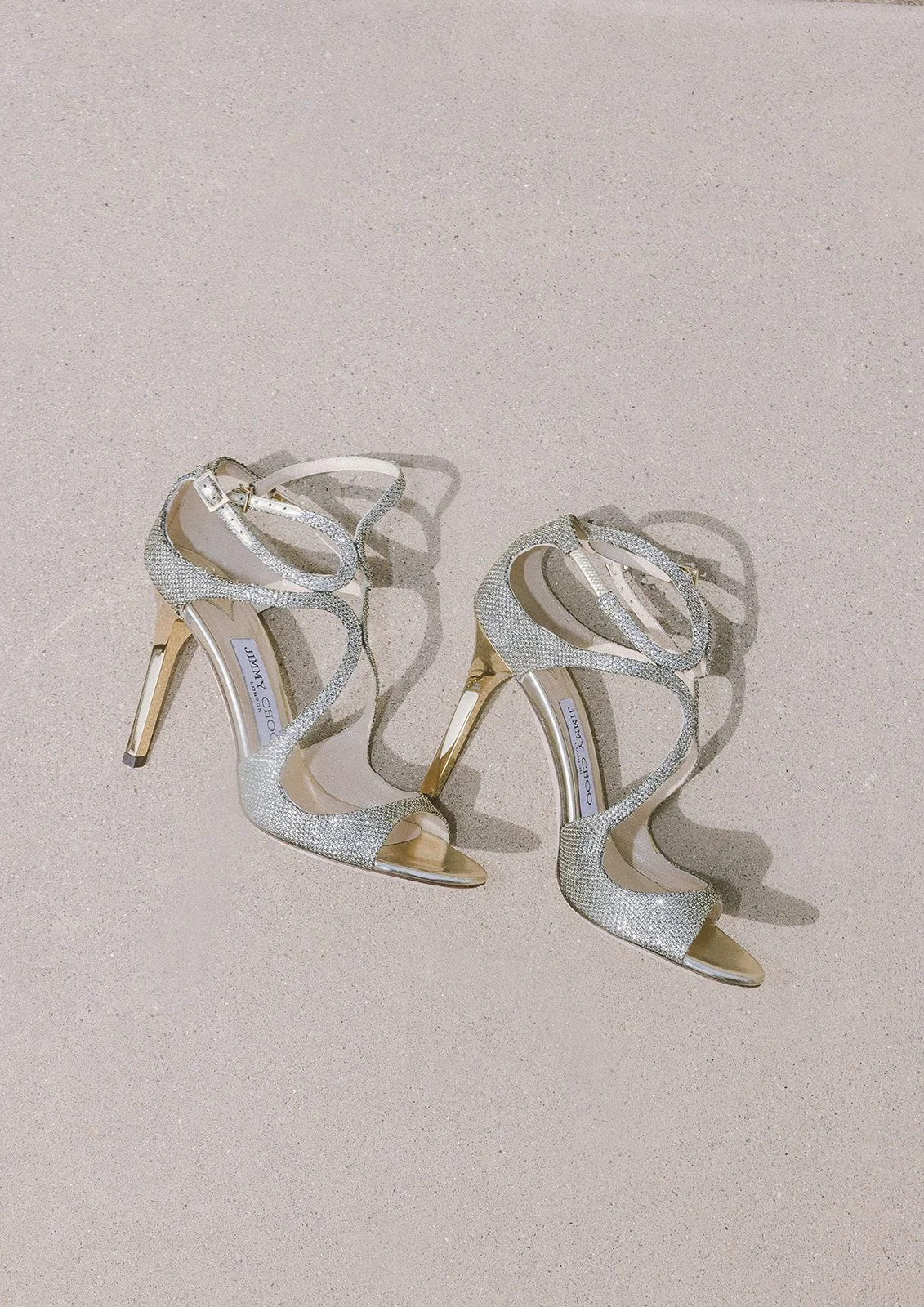 Pair of elegant high-heeled sandals with metallic straps on a sandy surface.