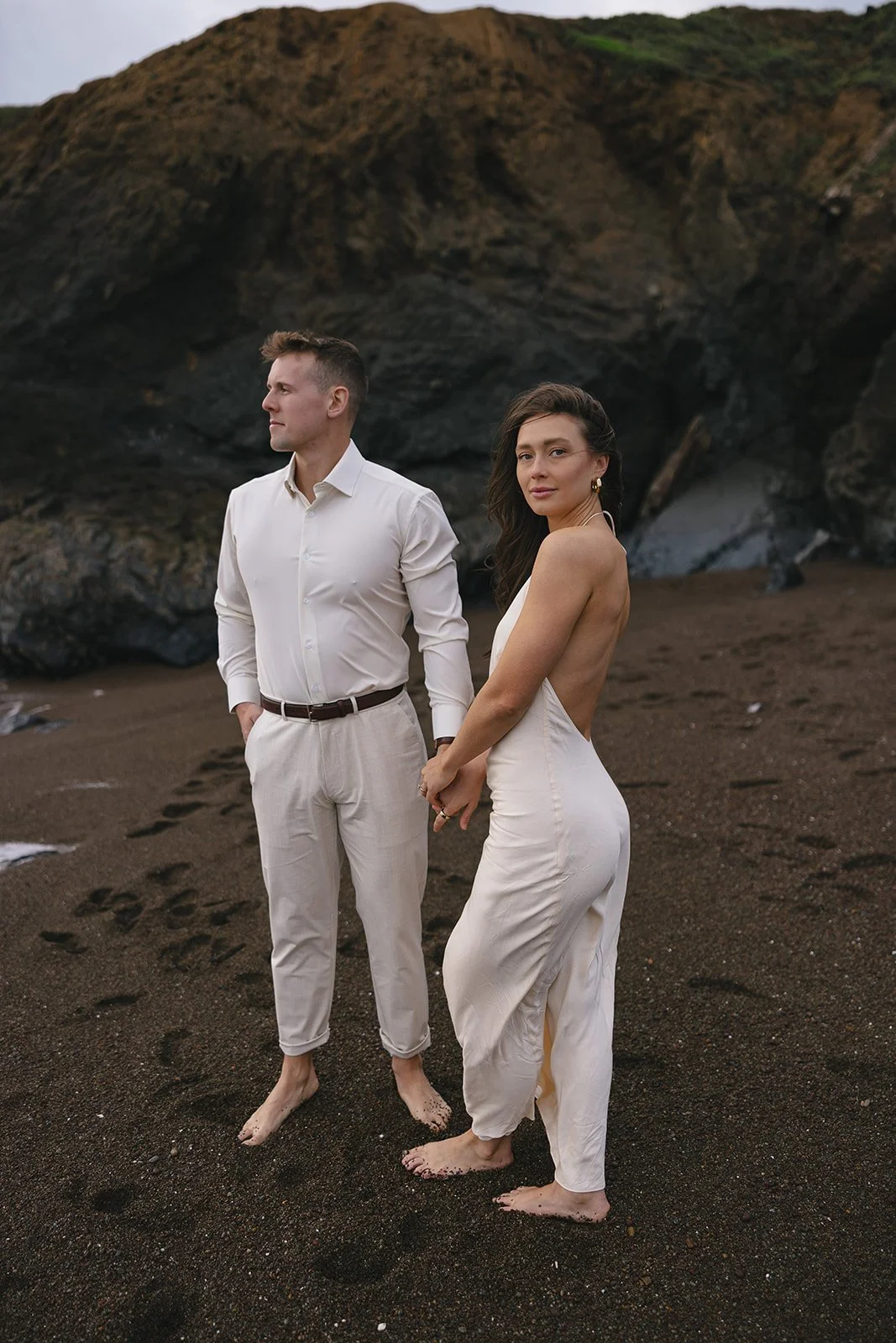 A man and woman stand barefoot on a sandy beach, holding hands and wearing elegant white outfits, with rocky cliffs in the background.