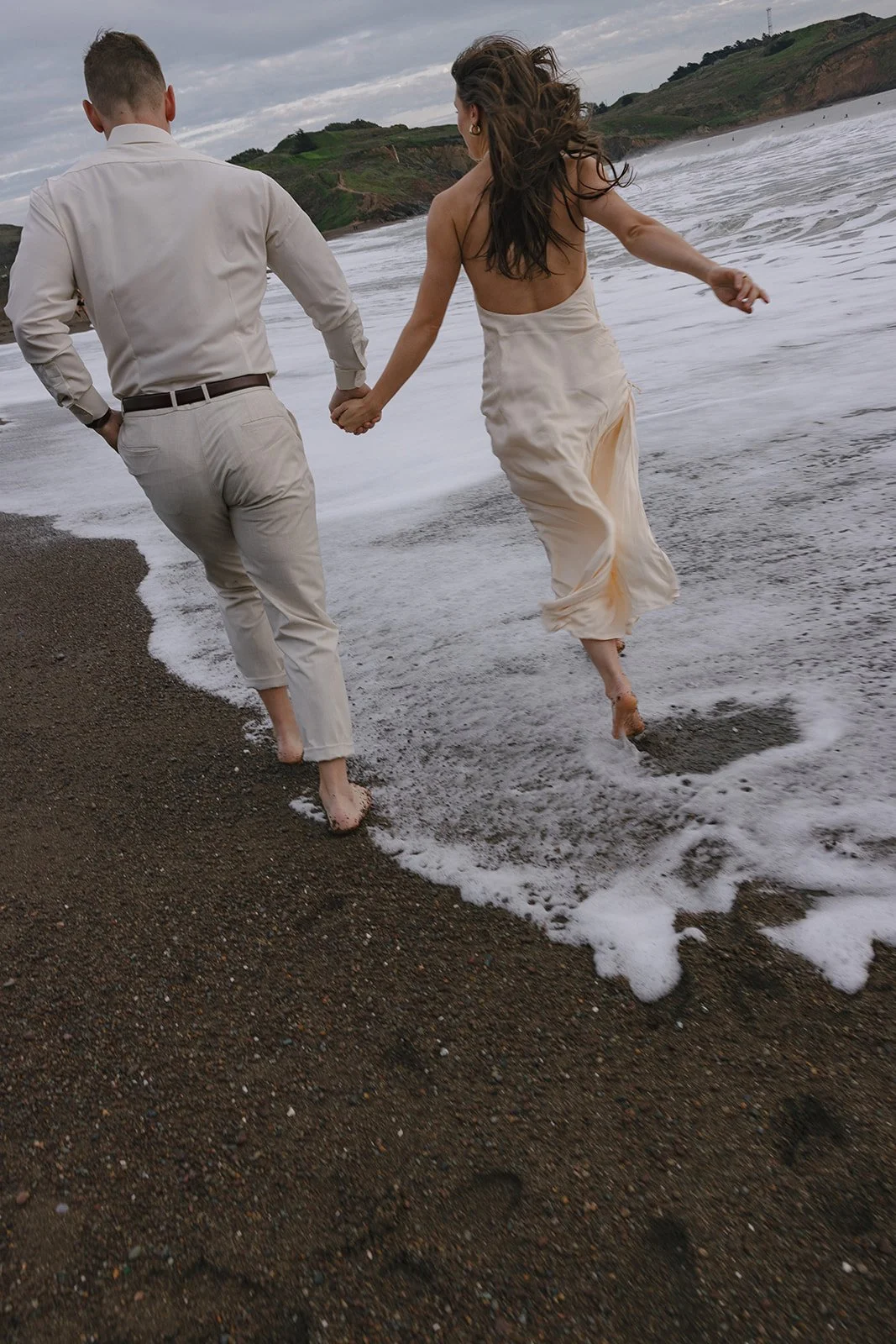 A couple holding hands, running barefoot along a beach with waves approaching, dressed in formal attire.