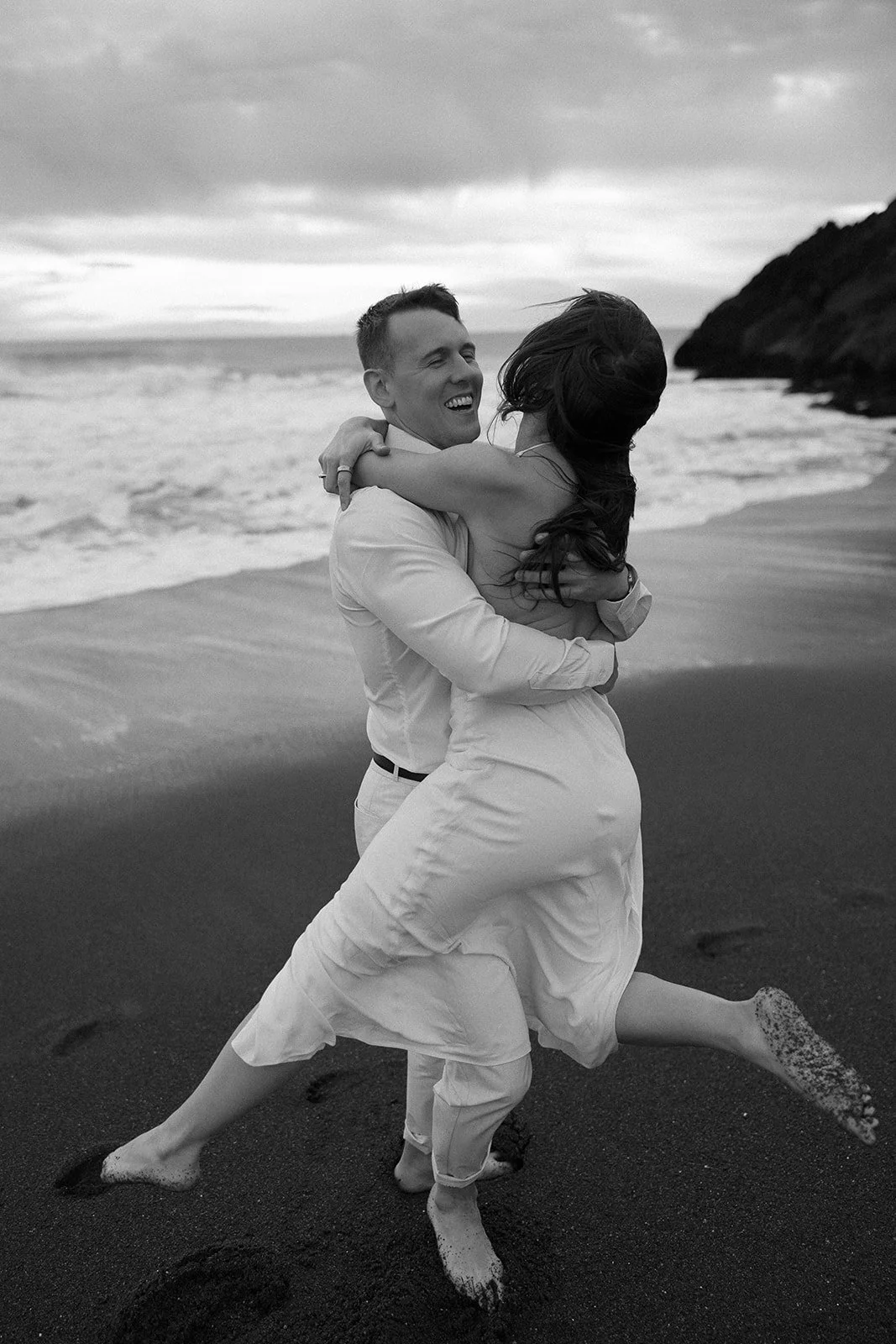 A joyful couple embracing on a sandy beach, with waves in the background. The woman is lifted in the man's arms, wearing a dress, and both are barefoot. The image is in black and white.