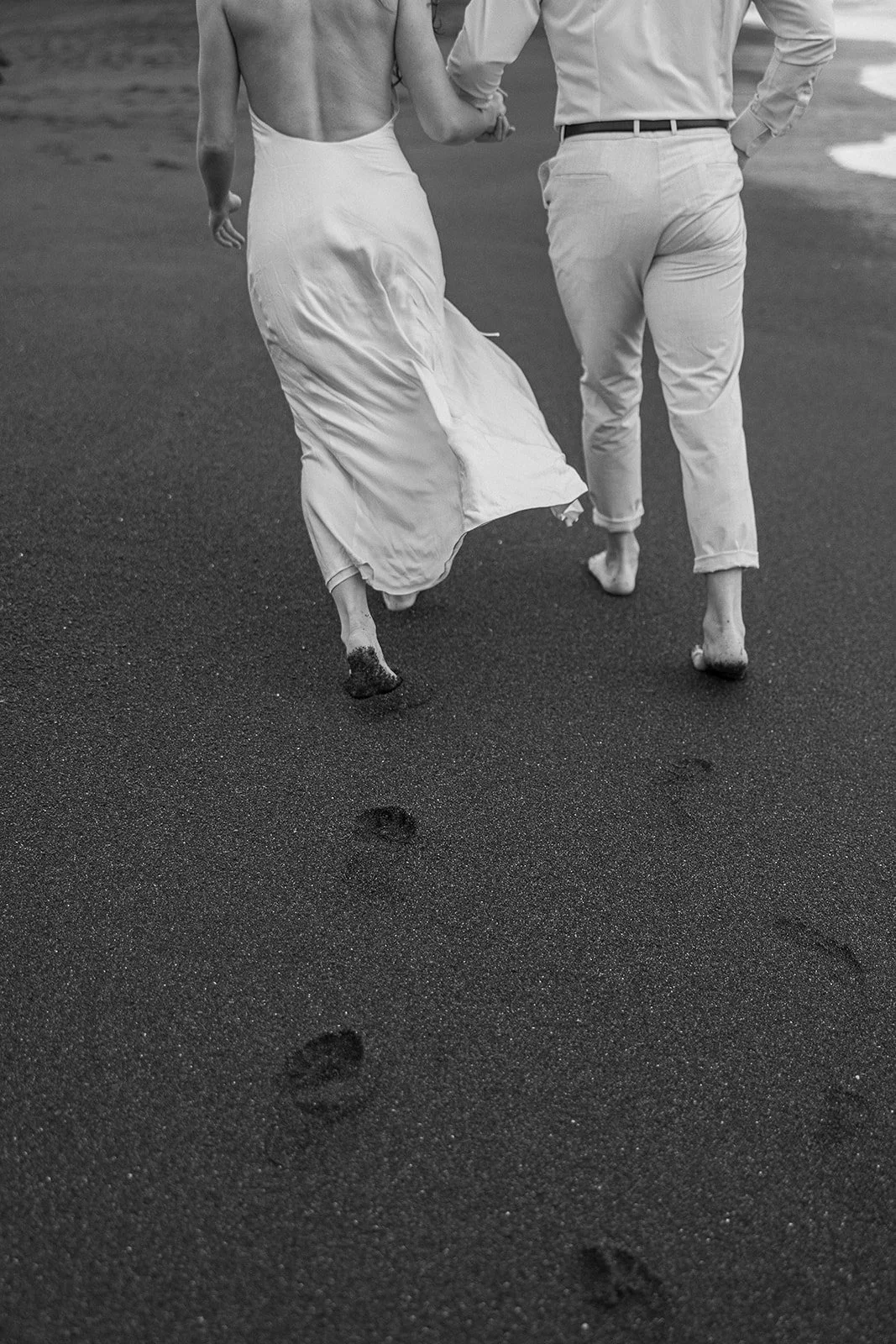 A black and white photo of a barefoot couple walking on sand, with the woman in a long dress and the man in light pants and a shirt, leaving footprints behind.