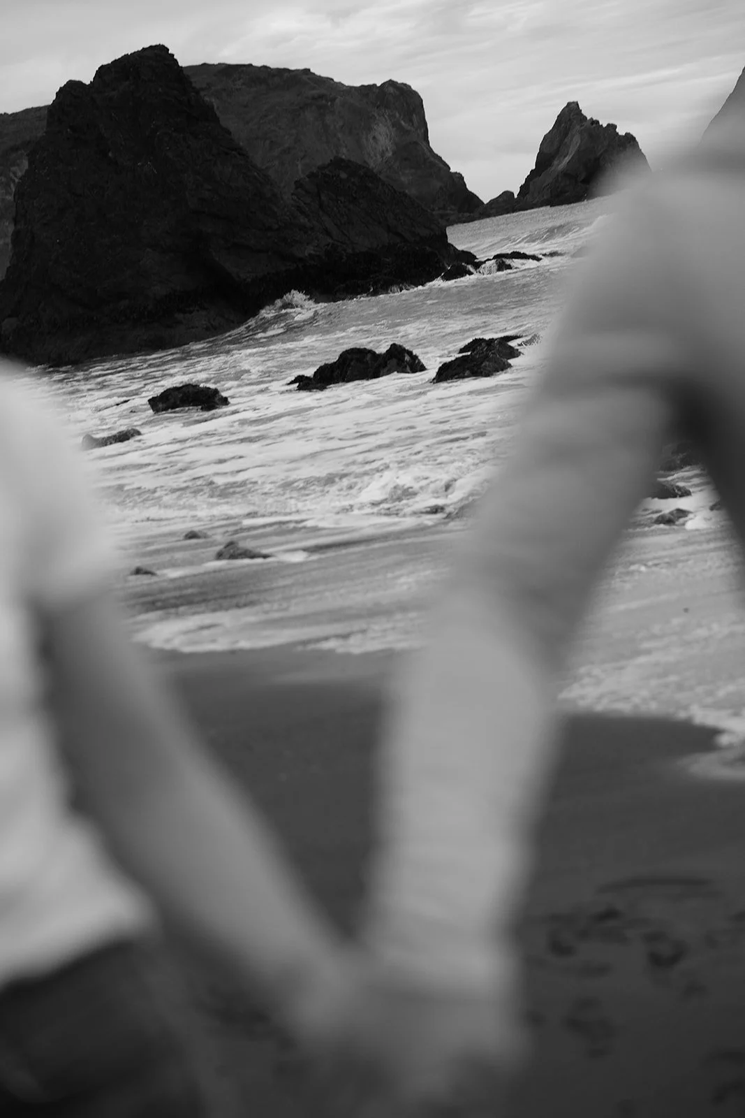 Black and white photo of a rocky coastline with waves and two out-of-focus people holding hands in the foreground.