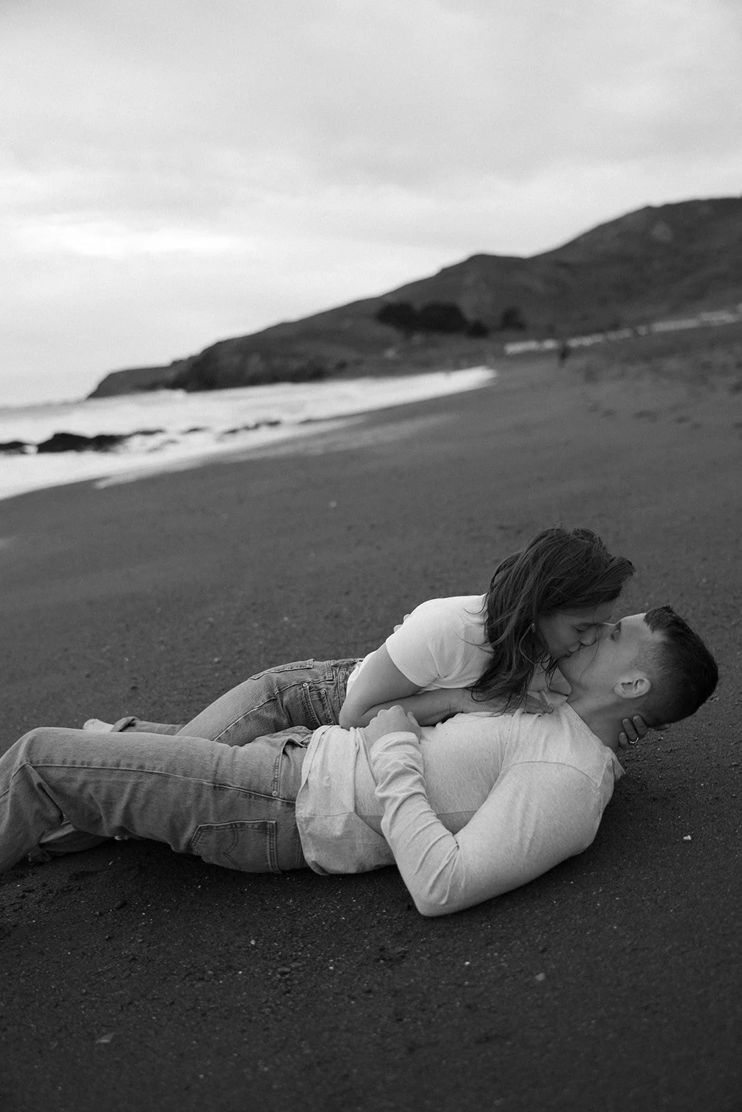 Black and white image of a couple kissing on a beach, with hills in the background.