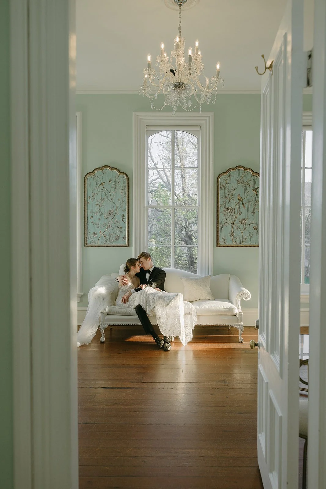 Bride and groom kissing on a white sofa in an elegant room with a chandelier and large window.