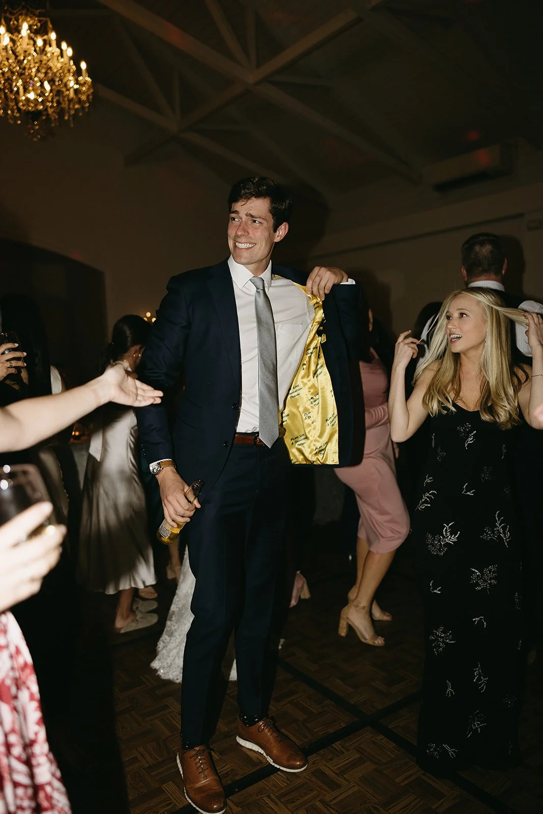 People dancing at a party, with a man in a suit holding a beer bottle and showing the inside lining of his jacket, while others enjoy the event.