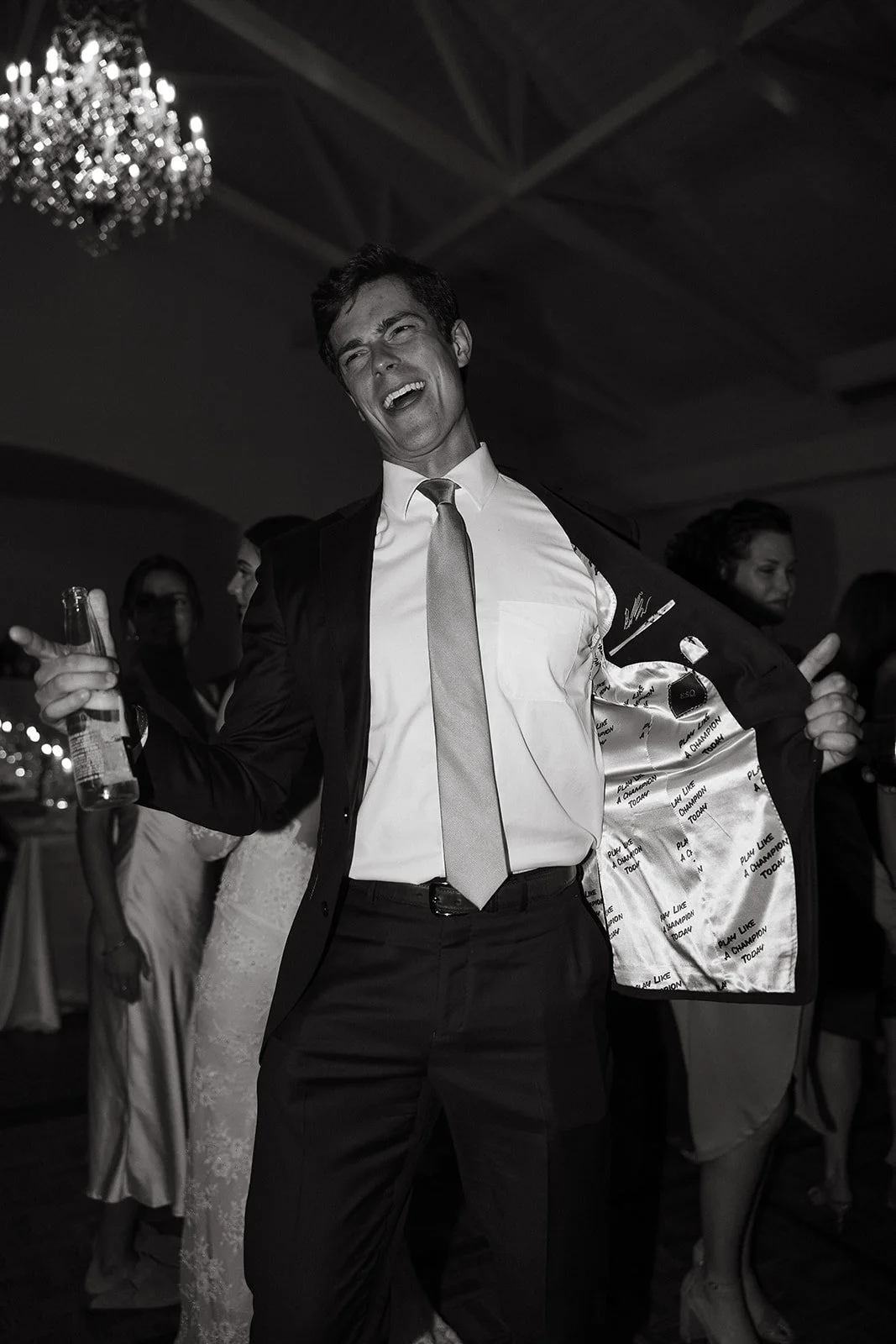 Man in a suit smiling and holding a drink at a party, with jacket lining visible. Other people in party attire in the background.