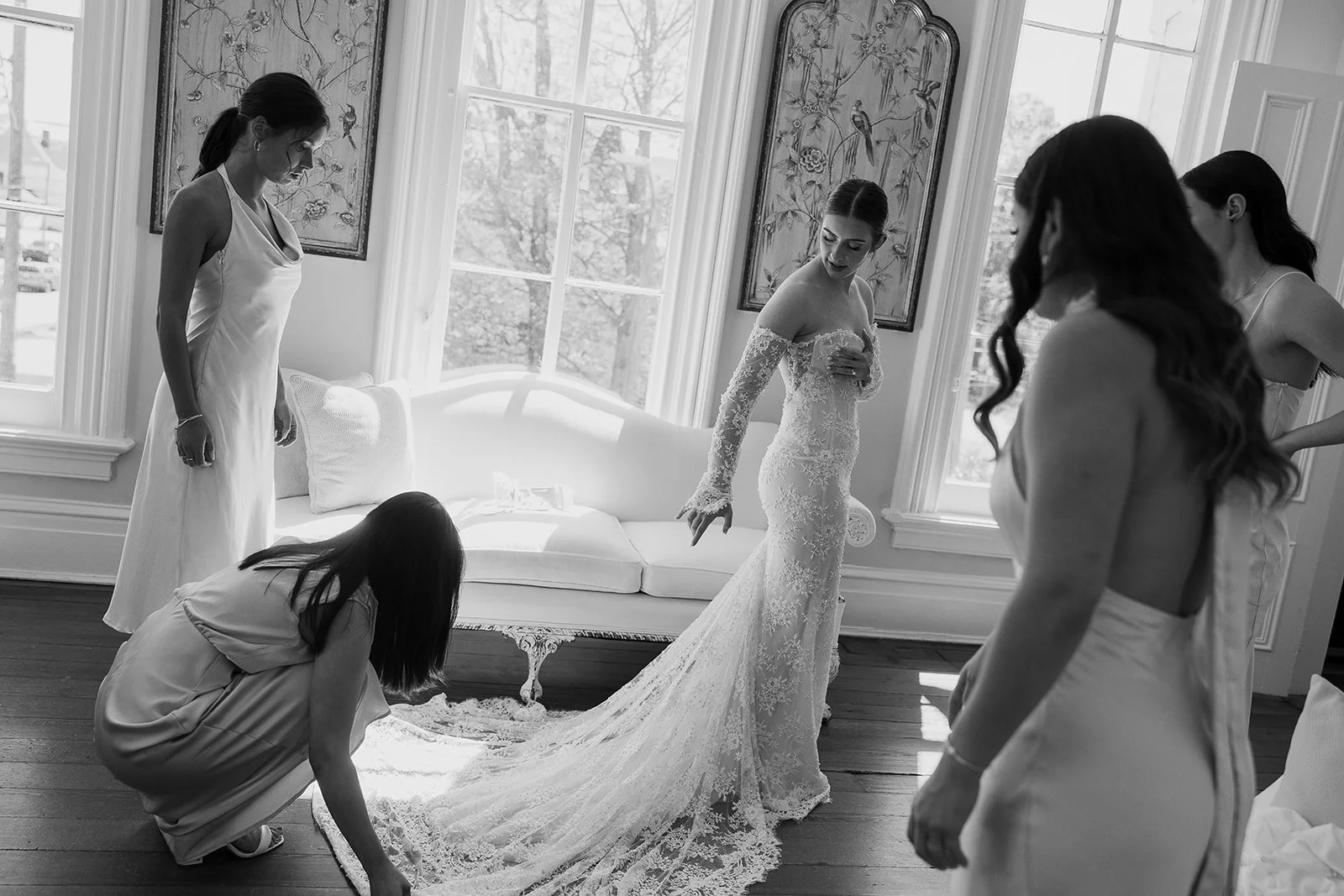 Bride wearing a wedding dress with lace sleeves surrounded by bridesmaids helping with her gown train in a sunlit room.