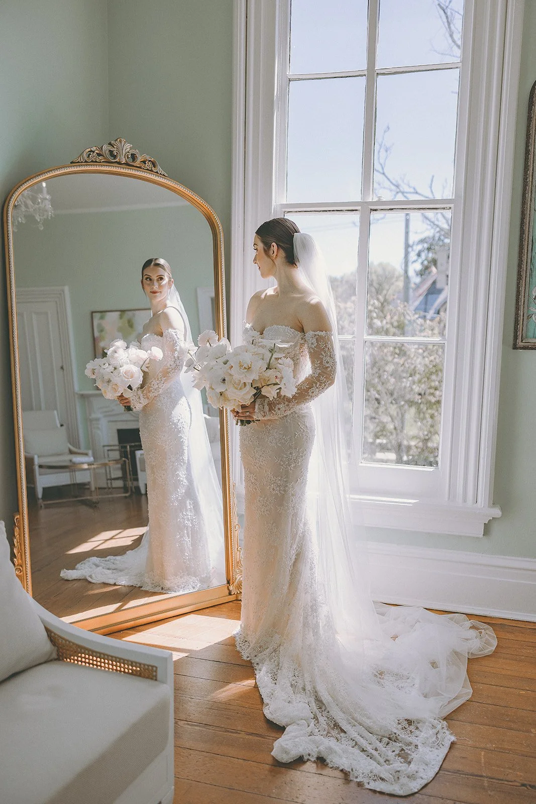 Bride in lace wedding dress holding bouquet, standing near window, reflected in ornate mirror.