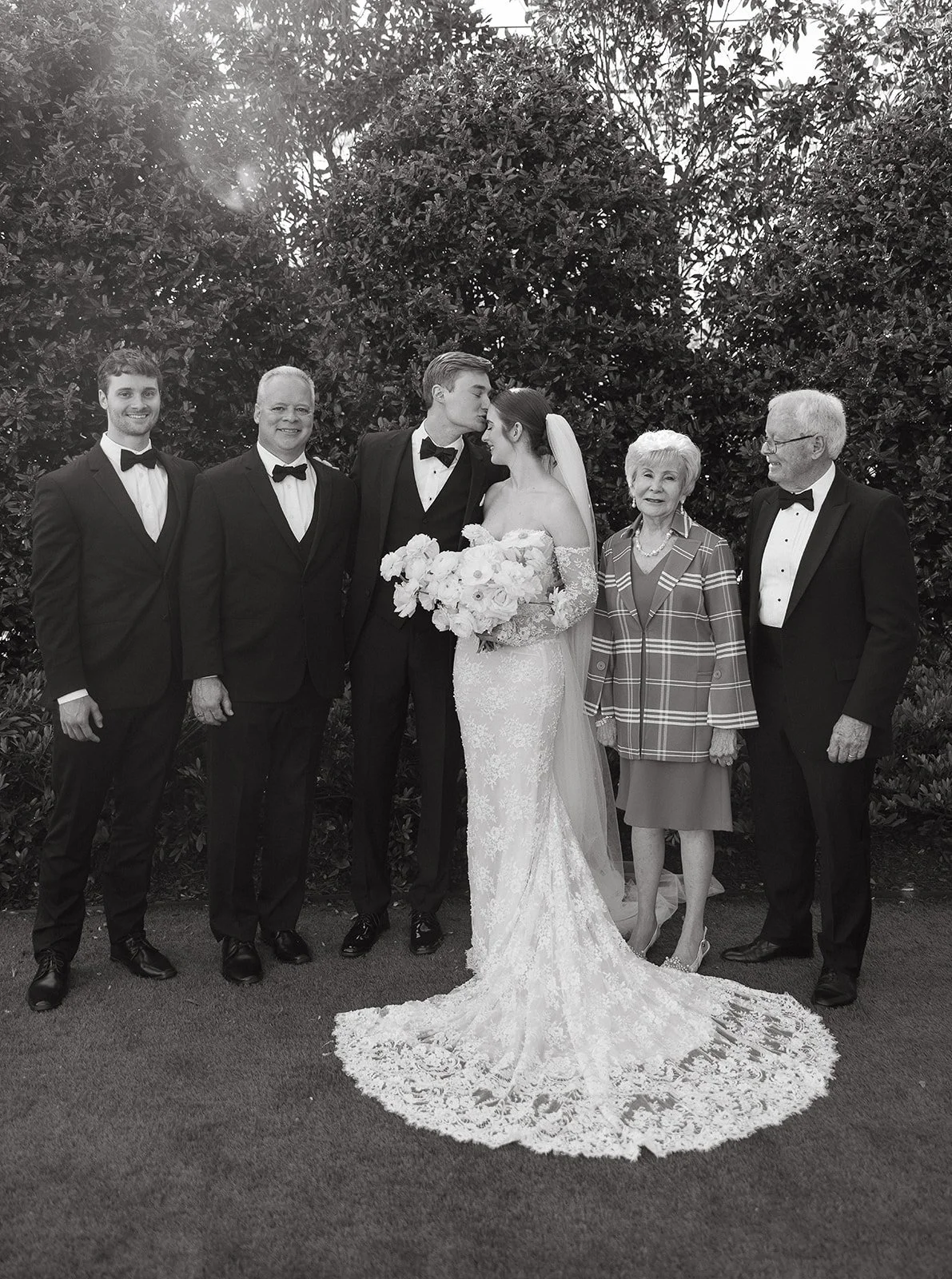 Black and white photo of a wedding group with a bride and groom kissing, surrounded by family members wearing formal attire, standing outdoors with leafy bushes in the background.