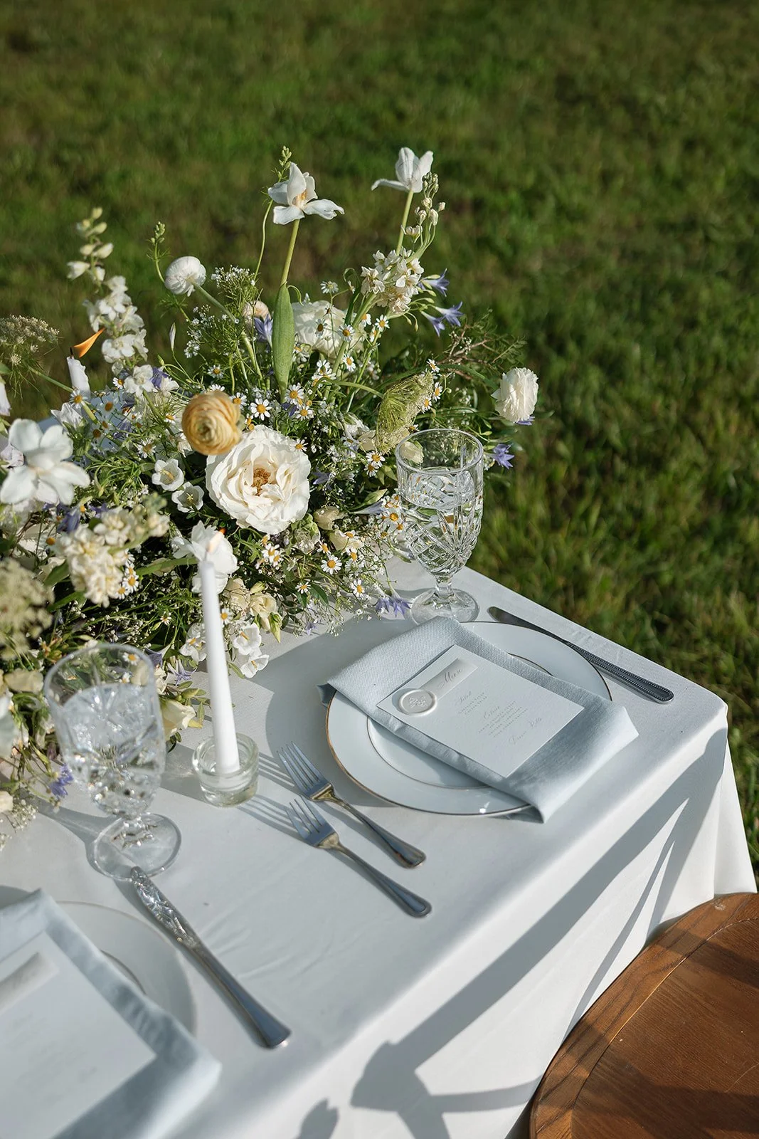 Elegant outdoor table setting with white tablecloth, glassware, cutlery, white plates, light blue napkins, menu cards, and a centerpiece of white and pastel flowers on grass.