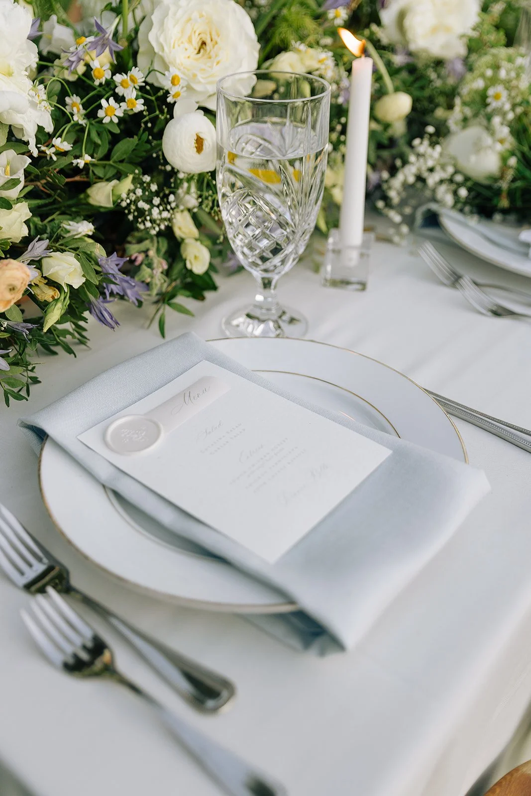 Elegant table setting with crystal glass, white plates, a menu on a napkin, and floral centerpiece featuring white and green flowers.