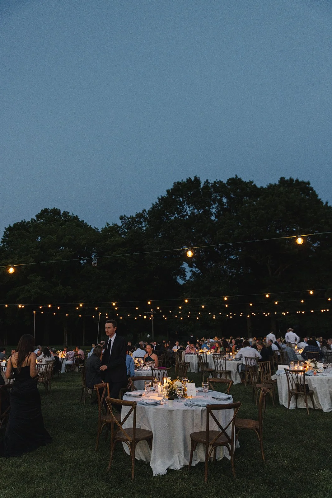 Evening outdoor wedding reception with string lights, round tables, and guests dressed in formal attire, set against a backdrop of trees.