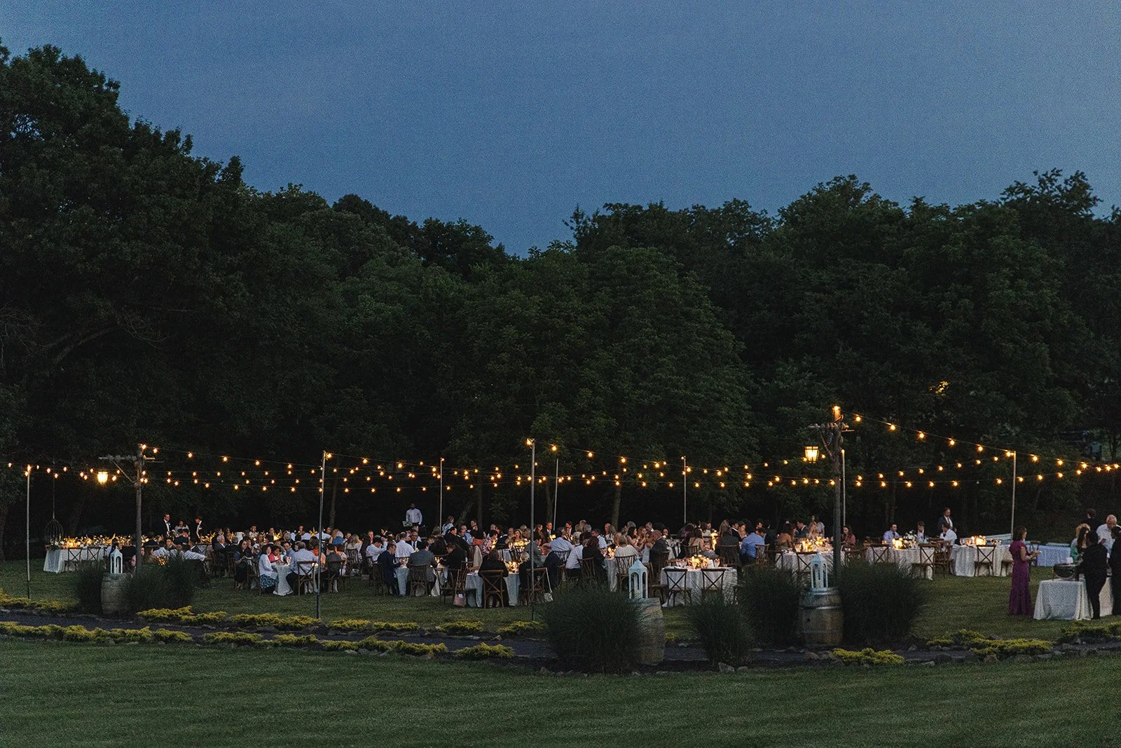 Outdoor evening event with guests seated at tables under string lights, surrounded by trees.