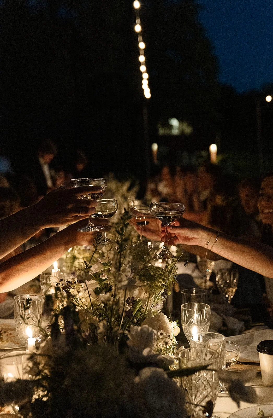 People raising glasses in a toast at an elegant outdoor evening event, with flower arrangements and candles on the table.