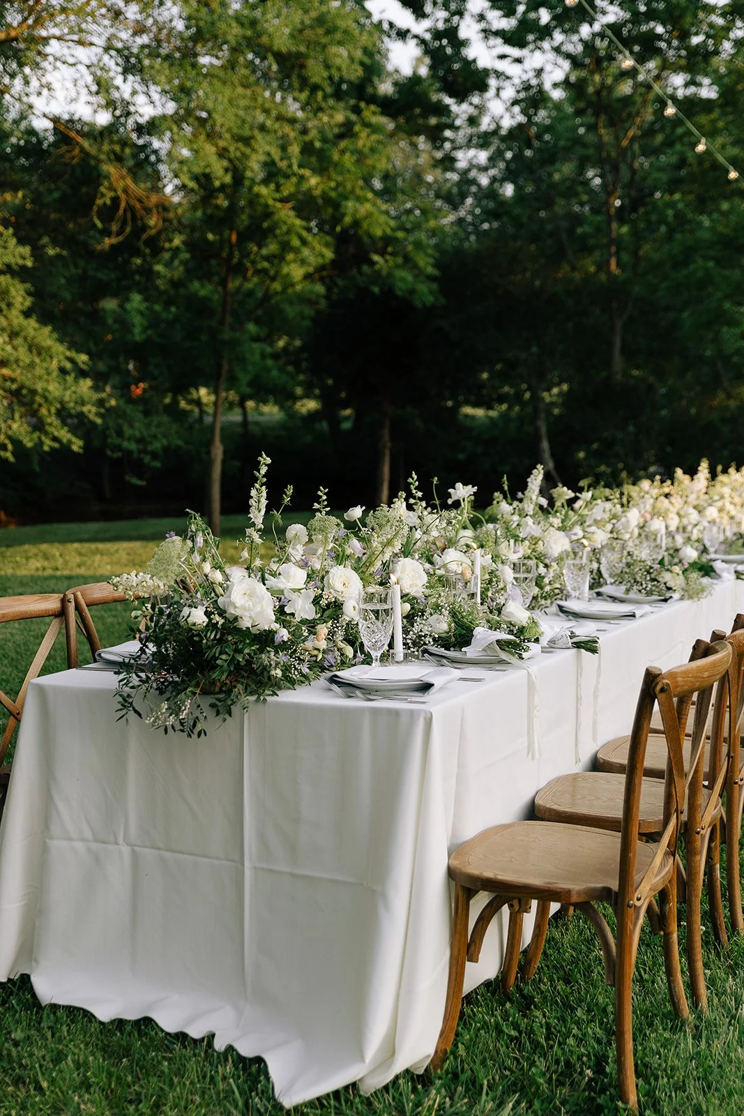 Outdoor dining table set with white tablecloth, wooden chairs, elegant floral centerpieces, and place settings in a garden setting.