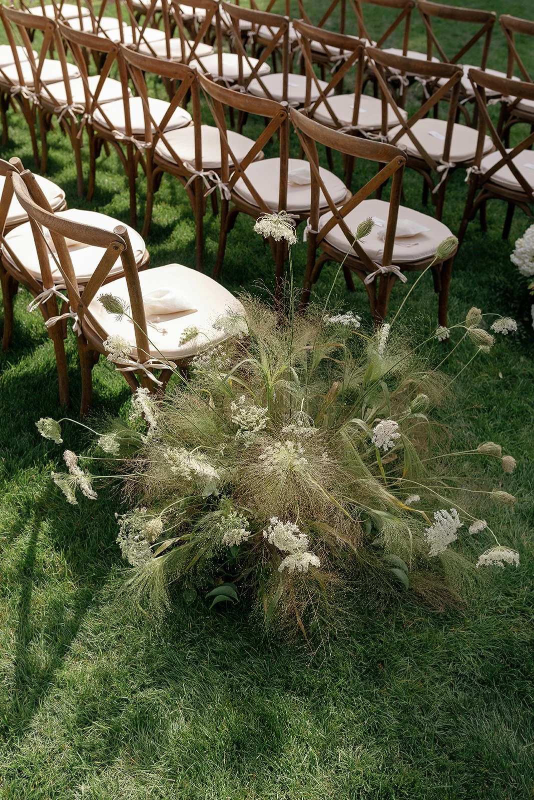 Rows of wooden chairs with white cushions, arranged on grass next to decorative wildflowers.