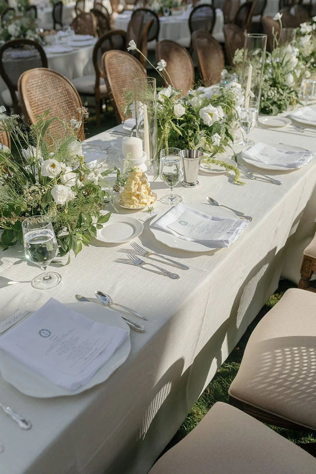 Elegant outdoor dining table setup with white tablecloth, floral arrangements, candles, and place settings, featuring glassware and menus.