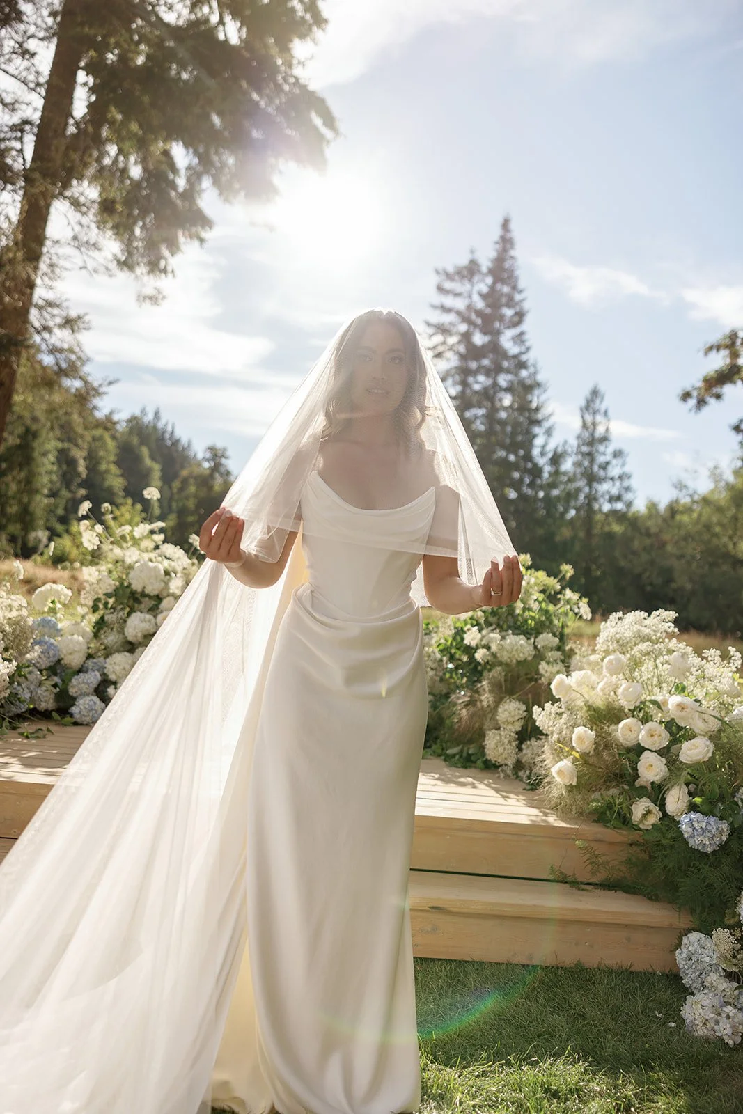 Bride in a white wedding dress and veil standing outdoors with floral arrangements and trees in the background.