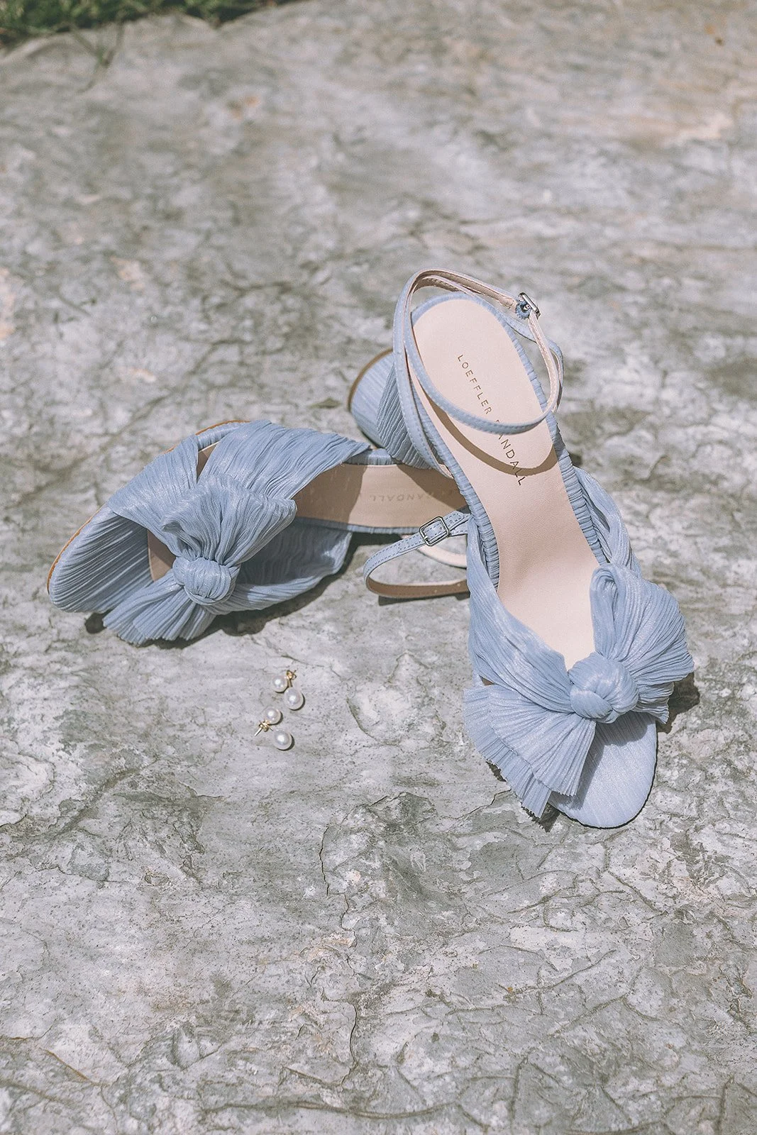 Light blue pleated sandals with a bow detail on a textured stone surface, next to a set of pearl earrings.