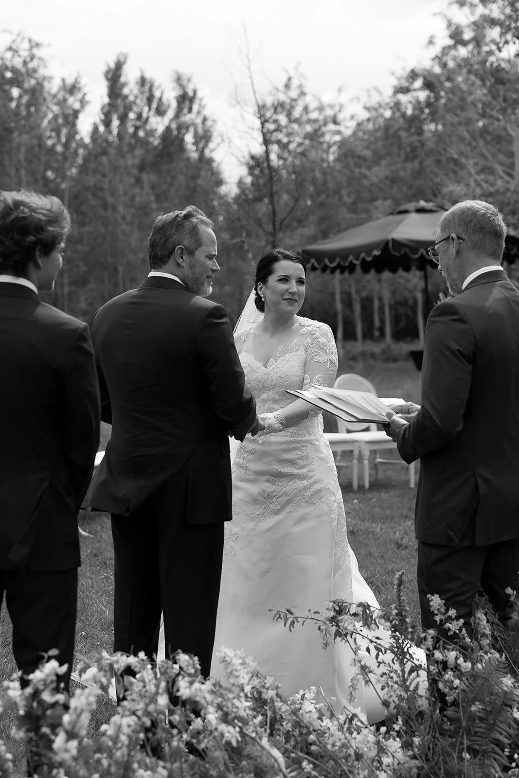 Black and white photo of an outdoor wedding ceremony, with a bride in a wedding dress and veil holding hands with a groom in a suit, officiant holding a document, and another person present. Trees and foliage are in the background.