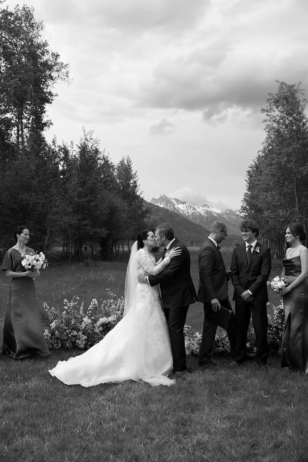 Bride and groom kissing during outdoor wedding ceremony with bridal party and mountains in background.
