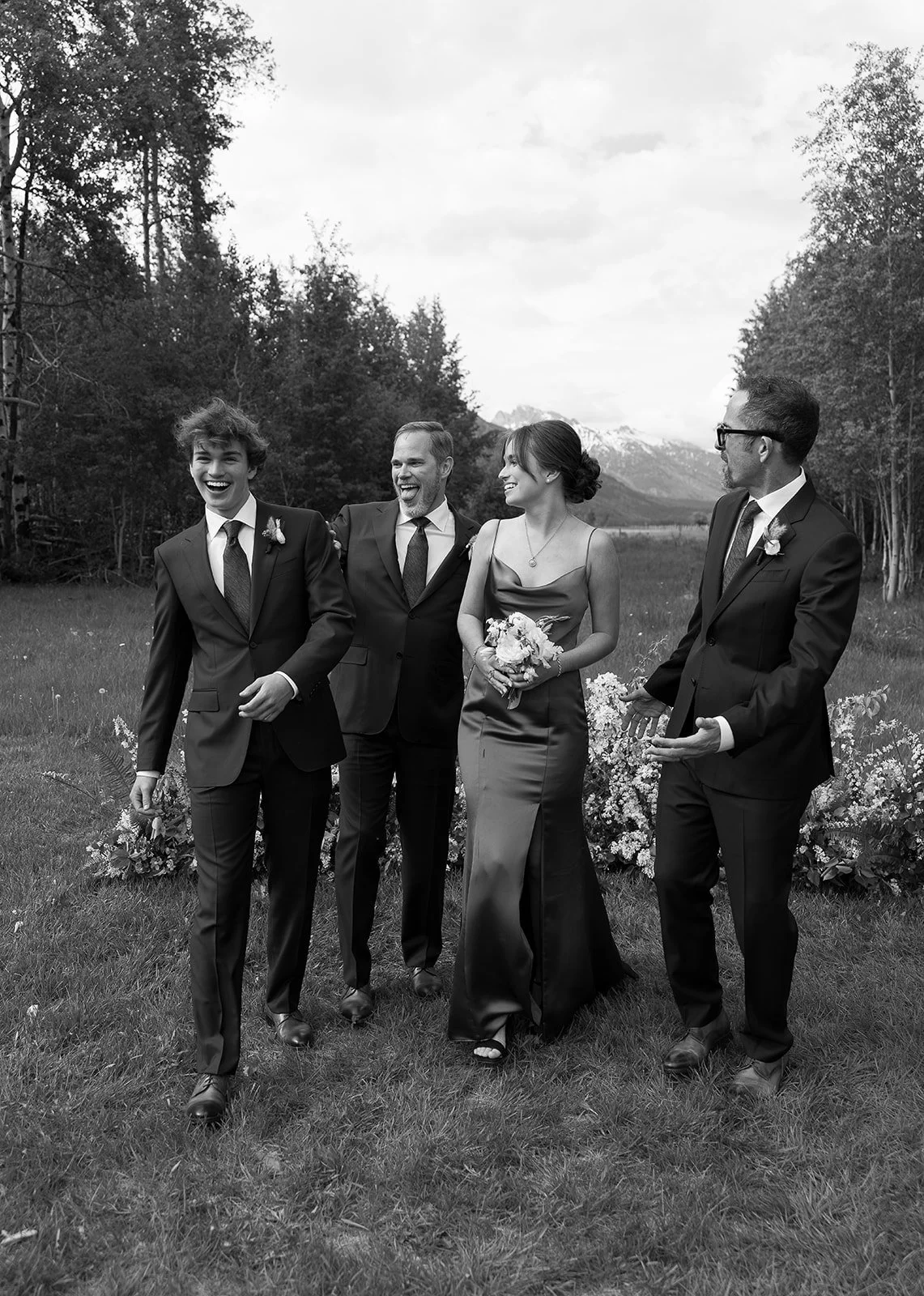 A black and white photo of four people dressed formally, smiling and walking outdoors on grass with trees and mountains in the background.