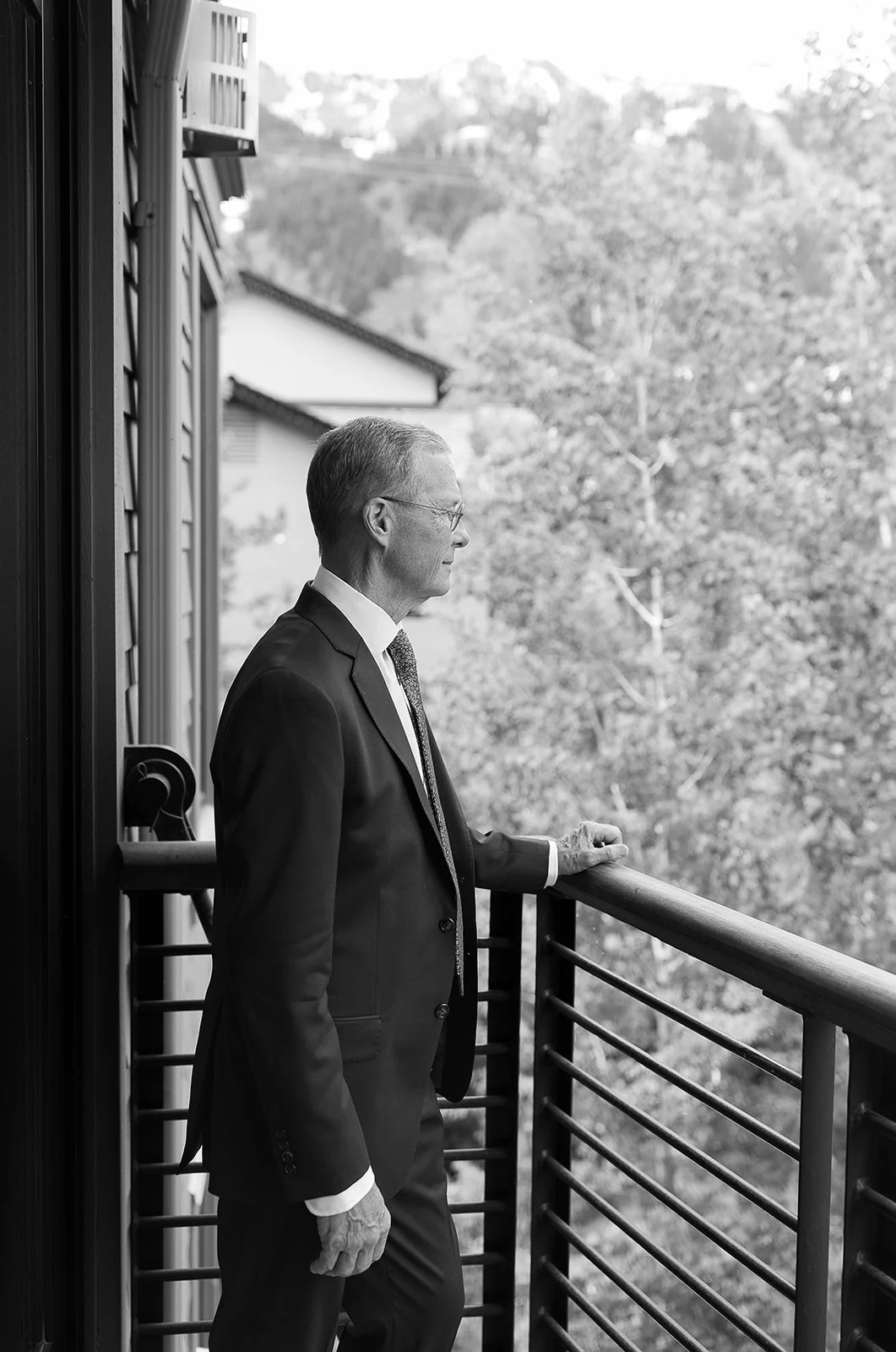 Man in a suit standing on a balcony overlooking trees.