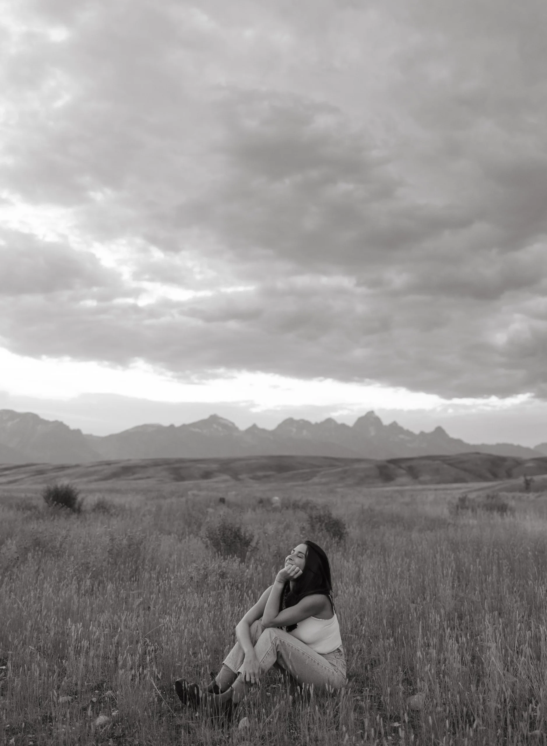 Black and white photo of a woman sitting in a field with mountains in the background.