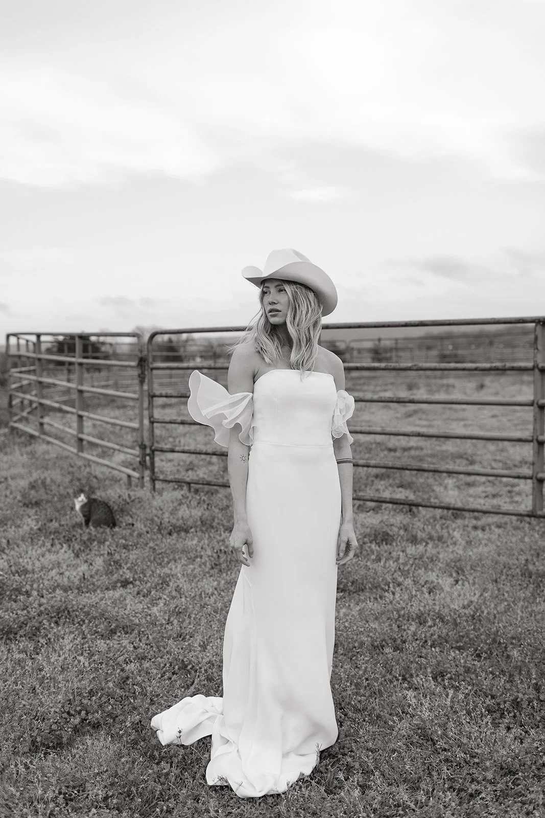 Woman in white wedding dress and cowboy hat standing in grassy field with metal fence, black and white photo.